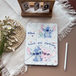 Puede incluir: Funda blanca para tableta con los personajes de Stitch y Angel y el texto "Love you forever". La funda tiene un diseño plegable e incluye un lápiz óptico blanco. Un calendario de madera muestra "19 de octubre."