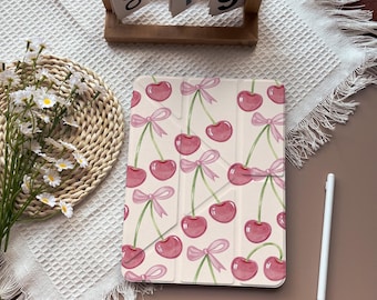 May include: A cream-colored tablet case featuring a pattern of pink cherries with green stems and pink bows. The case is open, revealing the interior. A white stylus and a wooden calendar are also visible.