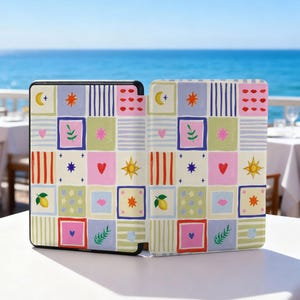 May include: A patterned tablet case with a colorful patchwork design. The design includes squares with various illustrations such as hearts, stars, moons, and lemons. The case is black with a white table and blue ocean in the background.