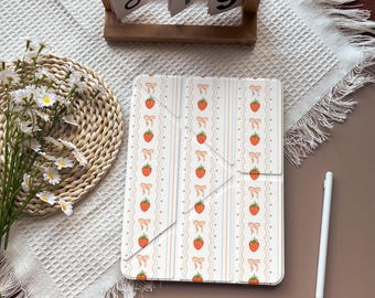 May include: A white tablet case with a repeating pattern of strawberries and bows. The design features vertical stripes in light blue and peach. A wooden calendar displays "Oct 19". A white stylus rests nearby.