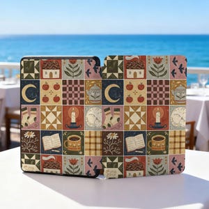 May include: A decorative case featuring a patchwork design with cozy, autumnal illustrations. The design includes houses, apples, candles, moons, socks, teacups, books, and floral elements in a palette of warm colors. The text "oh jess marie" is visible.