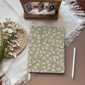 May include: A rectangular tablet case featuring a sage green background with a repeating pattern of small pink flowers and green stems, accompanied by a white stylus pen positioned to the right of the case on a flat surface.