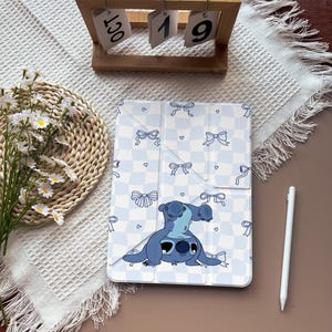 Peut inclure: Une housse de tablette bleu clair et blanche avec un personnage de dessin animé et un motif de nœuds. L'étui a un fond à carreaux bleu et blanc avec des nœuds et des cœurs bleus. Un stylet blanc est à côté de l'étui.