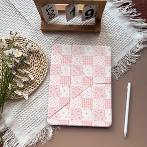 Peut inclure: Une coque de tablette rectangulaire présentant un motif à carreaux rose et blanc avec des carrés alternés d'imprimés floraux, de vichy et de pois. La coque a un design à trois volets et est placée à côté d'un stylet blanc sur une surface texturée.