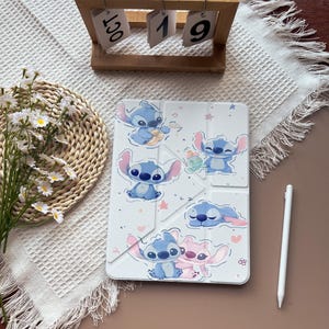 May include: White tablet case featuring blue cartoon characters with large ears. The case has a folding design and is decorated with hearts and stars. A white stylus is placed next to the case. A wooden calendar displays the date.