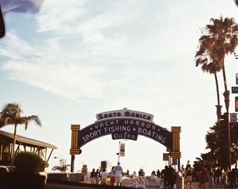 Peut inclure: Scène ensoleillée à Santa Monica, Californie. L'image présente une arche avec l'inscription "Santa Monica Yacht Harbor Sport Fishing Boating Cafes". Des palmiers et des personnes sont visibles sous un ciel bleu clair.