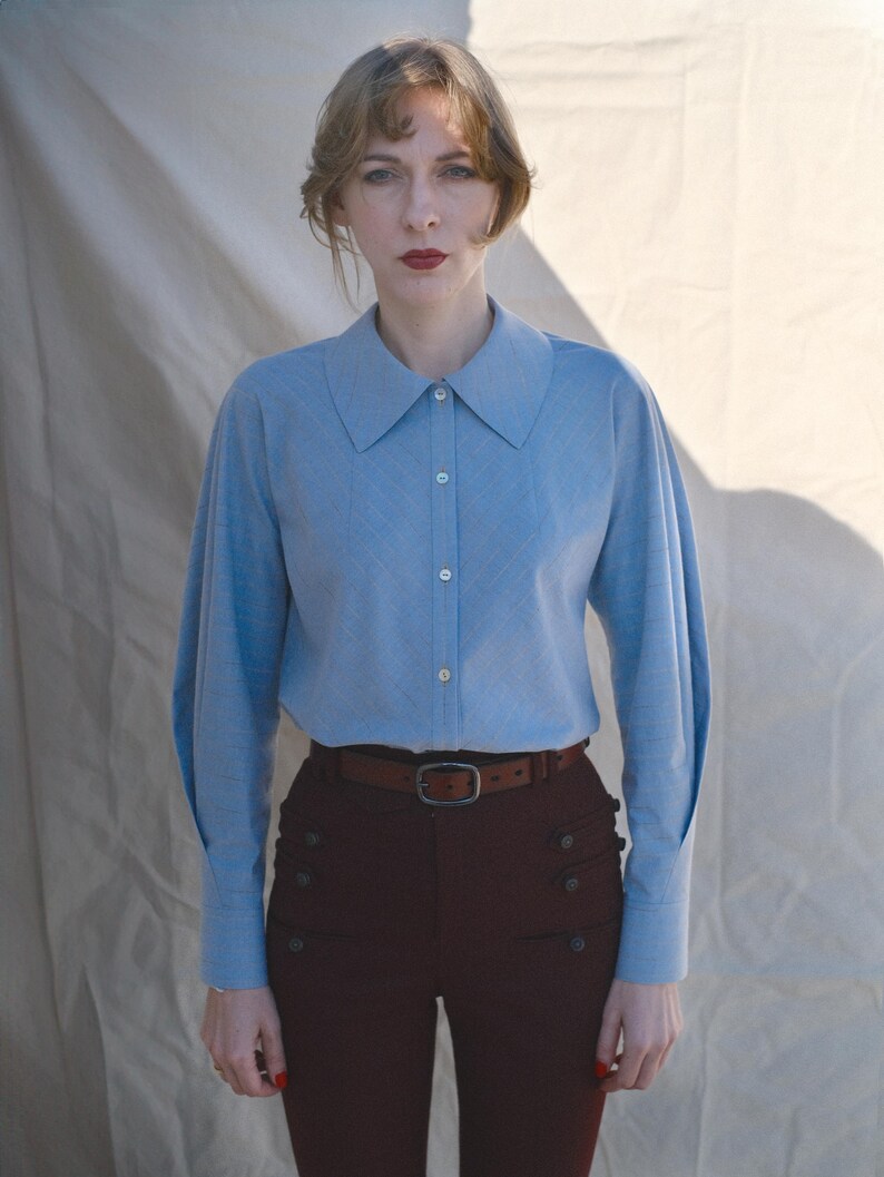 May include: A light blue long-sleeve button-down shirt with a pointed collar and full sleeves, paired with dark brown high-waisted trousers featuring two vertical rows of buttons and a brown leather belt with a rectangular metal buckle.
