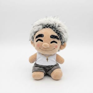 Può includere: Una bambola di peluche con pelle abbronzata, guance rosate e folte sopracciglia nere. La bambola ha capelli corti e appuntiti bianchi e neri, indossa una canottiera bianca con una collana a croce dorata e pantaloncini grigi con un sorriso.