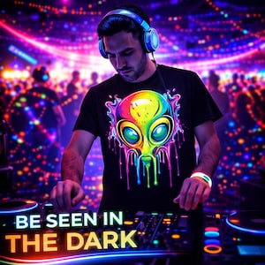 May include: A black t-shirt with a colorful alien graphic and the text "BE SEEN IN THE DARK". The image shows a person wearing blue headphones and a glowing wristband, operating DJ equipment in a club setting.