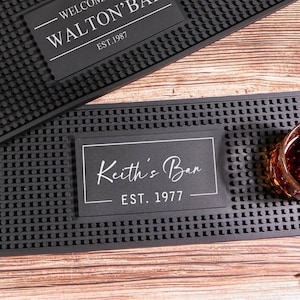 Puede incluir: Dos alfombrillas de bar negras con el texto "WELCOME TO WALTON'BAR EST. 1987" y "Keith's Bar EST. 1977". Una botella de whisky, dos botellas de cerveza y dos vasos sobre una superficie de madera.