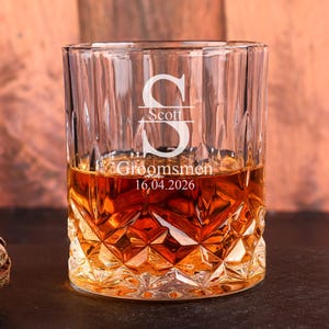 Puede incluir: Un vaso de whisky personalizado, transparente, con base tallada en diamante y ranuras verticales. El vaso está grabado con la letra "S", el nombre "Scott", la palabra "Groomsmen" y la fecha "16.04.2026". Contiene líquido ámbar.