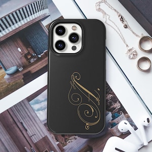 May include: Black phone case featuring a gold script initial design. The case has a protective bumper around the camera lenses. The background includes jewellery, a magazine, and wireless earbuds.