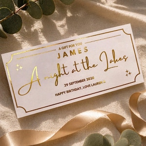 May include: A cream-coloured gift voucher with gold foil detailing. The text reads "A Gift For You, James, A Night at the Lakes, 29 September 2026, Happy Birthday, Love Lauren". The card is rectangular with decorative edges.