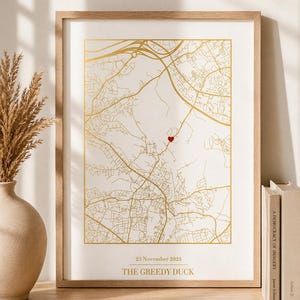 May include: A framed map print with gold lines on a cream background. A red heart marks a location. The text "23 November 2024" and "THE GREEDY DUCK" are printed below the map. The frame is light brown.