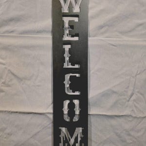 May include: A dark grey metal sign with the word "WELCOME" cut out vertically. The letters are white. The sign is designed for display, suitable for a door or wall. The sign is made of metal.
