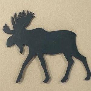 May include: A dark teal metal moose silhouette with large antlers. The moose is depicted in a walking pose, with its legs extended. The background is a light tan color.