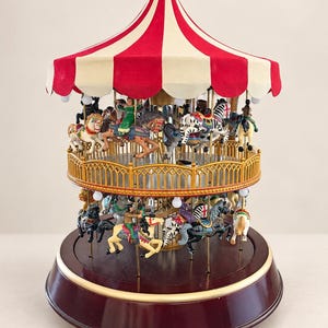 May include: A two-tiered carousel music box featuring a red and white striped canopy. The structure includes gold-colored railings and multiple hand-painted horses and figures on poles. The base is a dark maroon color with a gold trim accent.