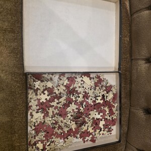 May include: An open rectangular box filled with puzzle pieces. The pieces are a mix of burgundy and cream colors, with text visible on some. The box is black with a white interior.