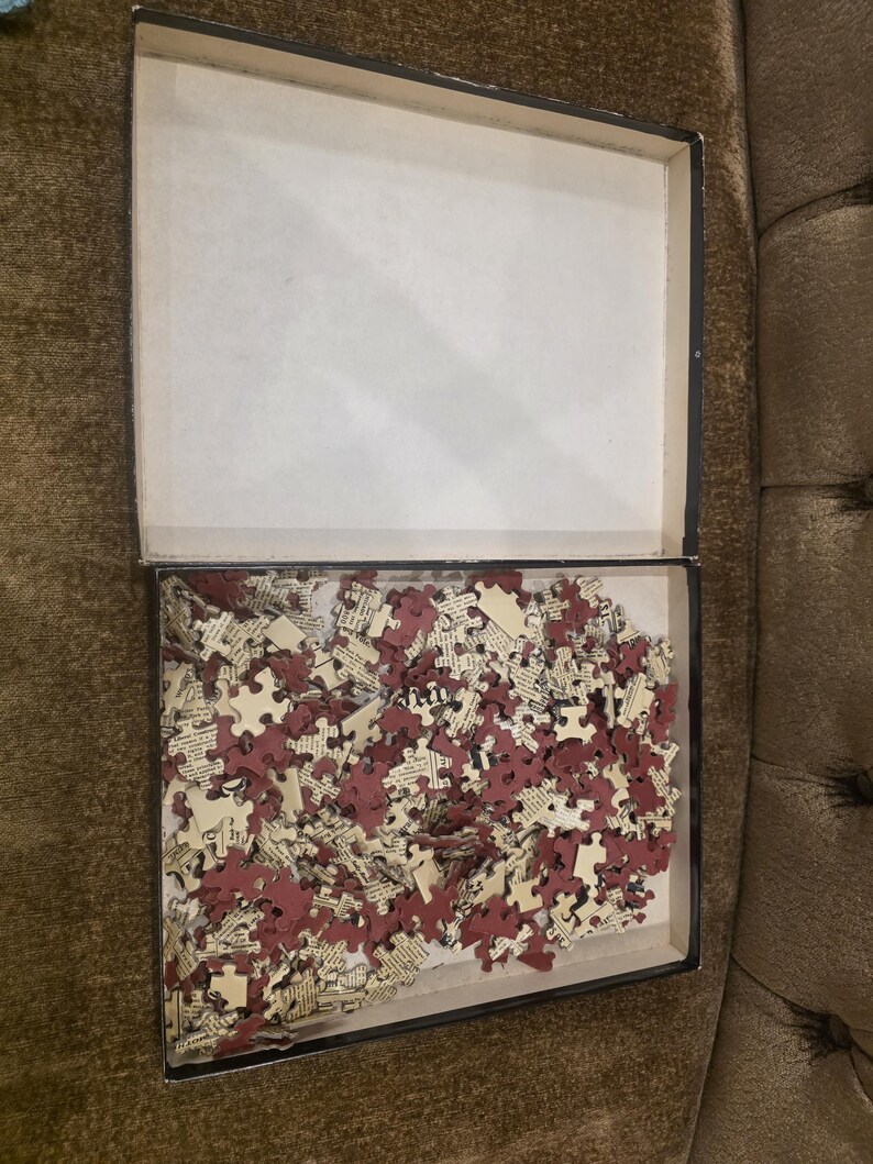 May include: An open rectangular box filled with puzzle pieces. The pieces are a mix of burgundy and cream colors, with text visible on some. The box is black with a white interior.
