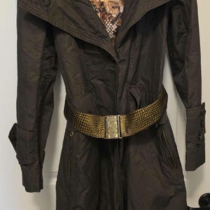 Puede incluir: Un abrigo tipo trench marrón oscuro con cinturón y detalles dorados. El abrigo tiene cuello, mangas largas y forro estampado. El cinturón tiene una hebilla decorativa. El abrigo está colgado en una puerta blanca.