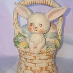 May include: A ceramic Easter bunny figurine in a woven basket. The bunny has long ears, rosy cheeks, and is surrounded by painted flowers and eggs. The basket is beige with a curved handle, perfect for Easter decor.