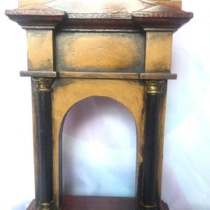 May include: A wooden decorative archway with a dark brown base and pillars. The arch is painted in shades of gold and brown, with black accents on the pillars and top. Three black cylindrical shapes are on top of the arch.