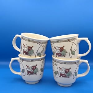 May include: Four white ceramic mugs with a floral design featuring red, green, and white flowers and leaves. The mugs have a cream-colored border and a curved handle. The background is a solid blue.