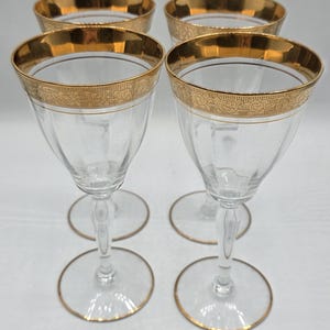 Può includere: Quattro calici da vino trasparenti con bordi e basi dorate. I bordi presentano un design dorato ornato. I calici sono disposti su una superficie bianca, mostrando la loro forma elegante e i dettagli.