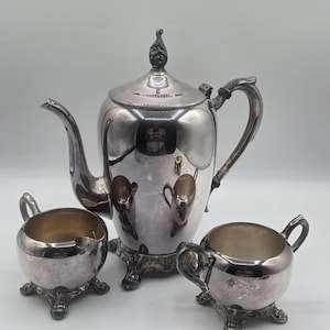Op de afbeelding: Een zilveren theeservies, bestaande uit een theepot, een melkkan en een suikerpot. De theepot heeft een gebogen tuit en een decoratieve knop. De set is gemaakt van glanzend metaal en staat op een lichte achtergrond.