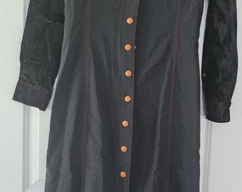 May include: A long, dark gray dress with a button-down front and a black velvet collar and sleeve accents. The dress features red stitching and copper-colored buttons, creating a striking contrast. The dress is a long, elegant style.