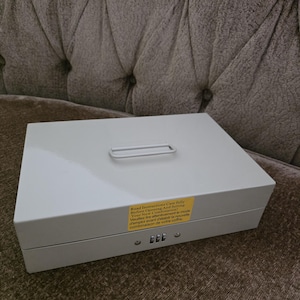 May include: A white metal cash box with a silver handle and a combination lock. A yellow sticker with instructions is affixed to the top. The box is rectangular and sits on a brown surface.