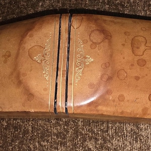 Puede incluir: Un estuche rectangular de color marrón claro para gafas con tapa abisagrada. El estuche tiene un aspecto desgastado y moteado con manchas más oscuras y detalles decorativos dorados. El estuche está dividido en dos secciones por una línea vertical.