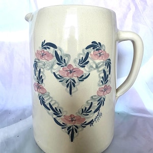 May include: Cream-coloured ceramic jug with a heart-shaped floral design. The heart features pink flowers and blue leaves. The jug has a handle and a spout. The artist's signature and the year 1994 are visible on the side.