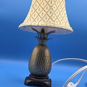 May include: A decorative table lamp with a pineapple-shaped base and a patterned lampshade. The base is bronze-colored with a textured design, and the shade is cream-colored with a diamond pattern. The lamp sits on a black base and has a white power cord.