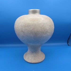 Puede incluir: Un jarrón decorativo con un cuerpo bulboso y una base delgada. El jarrón tiene un acabado texturizado de color gris claro con un borde plateado. El jarrón es una pieza única de decoración del hogar.