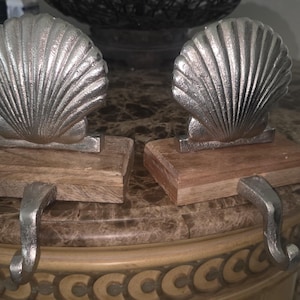 May include: Two silver-colored seashell stocking holders. Each shell is mounted on a rectangular wooden base with a curved metal hook. The holders are designed to hang stockings during the holidays.