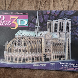May include: A box for a 3D puzzle of Notre Dame Cathedral. The box features a detailed image of the cathedral in shades of beige, gray, and green. The text "Super Challenging" and "Puzz 3D" are visible.