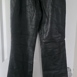 Puede incluir: Pantalones de cuero negro con cierre de botón y cremallera. Los pantalones cuelgan de una percha blanca contra una puerta blanca. Los pantalones tienen un ligero acampanado en la parte inferior.