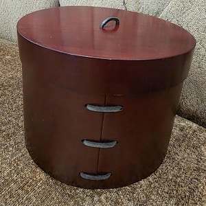 May include: A round, dark brown hat box with a matching lid. The lid has a small, curved handle. The box has three decorative, stitched accents on the side. The box is sitting on a textured, tan surface.