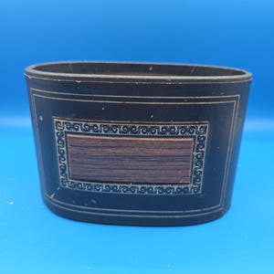 Könnte beinhalten: Ein dunkelblaues, oval geformtes Gefäß mit einer dekorativen rechteckigen Tafel. Die Tafel hat eine braune Holzmaserung in der Mitte, die von einer detaillierten, verzierten Bordüre umrahmt wird. Der Behälter steht vor einem blauen Hintergrund.