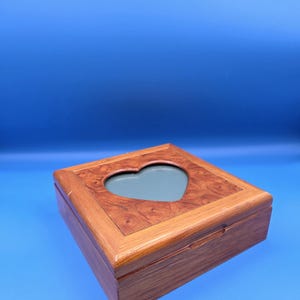 May include: A wooden jewelry box with a heart-shaped cutout on the lid. The box is rectangular with a light brown finish and a darker wood grain pattern on the top. The heart cutout reveals a gray interior.