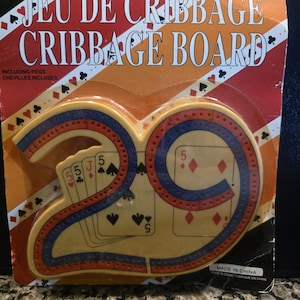 May include: A new cribbage board game in its original packaging. The board is shaped like the number 29, with red and blue accents. The packaging includes the text "JEU DE CRIBBAGE CRIBBAGE BOARD". Includes pegs.