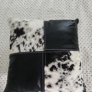 May include: Square decorative pillow with a cowhide and black leather design. The pillow features a patchwork pattern, with two sections of black leather and two sections of white and black cowhide. White stitching accents the seams.