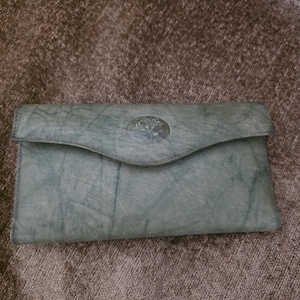 May include: A sage green leather wallet with a flap closure. The wallet features a decorative tree emblem on the front. The leather has a textured appearance. The wallet is rectangular in shape.