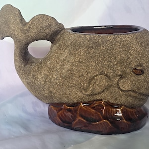 May include: A ceramic whale-shaped planter with a textured, speckled brown finish. The whale has a tail with a hole, a detailed face, and sits on a brown, wave-like base. The interior is a darker brown.