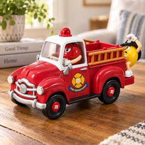 May include: A red and white toy fire truck featuring a red M&M character in the driver seat and a yellow M&M character on the back. The truck has a silver bumper, black wheels with red M logos, and a yellow emblem on the side that reads Fire Department.