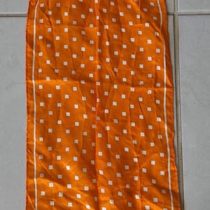 May include: A long, rectangular scarf in a vibrant orange color featuring a repeating pattern of small, white square dots. The scarf is framed by a thin white border that runs along all four edges, creating a clean and structured appearance.