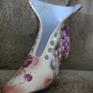 May include: A decorative porcelain shoe-shaped vase. The vase features a light blue interior and is painted with purple grapes and vines. Gold trim accents the edges and the small heel. The exterior is a cream color with pink and brown accents.