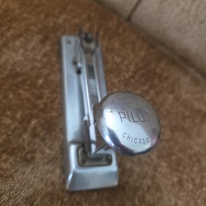 May include: A vintage silver-toned stapler with the word "PILOT" and "CHICAGO IL" engraved on the circular handle. The stapler is made of metal and has a rectangular body. The image is taken from a close-up angle.