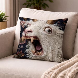 Puede incluir: Cojín decorativo cuadrado con un primer plano de una cabeza de cabra blanca y peluda con la boca abierta, revelando grandes dientes y una lengua rosada. El fondo es un estampado floral azul oscuro. El cojín está sobre un sofá beige.